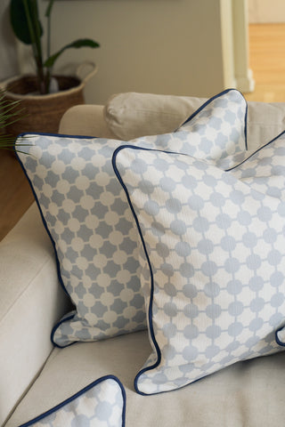 An Oversized Mod Dot Grid Pillow, featuring a hand-drawn exclusive pattern and dark blue piping, rests on a beige sofa. A plant in a woven basket adds to the modern Southern vibe.