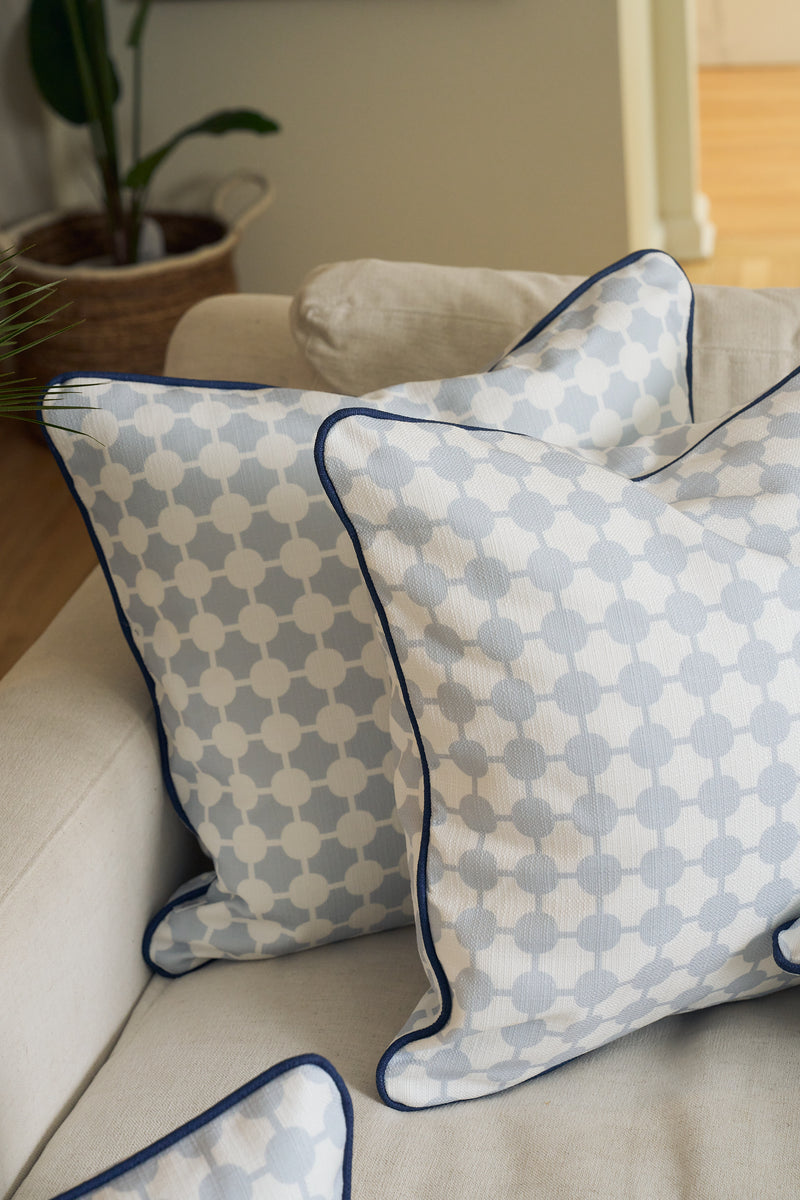 An Oversized Mod Dot Grid Pillow, featuring a hand-drawn exclusive pattern and dark blue piping, rests on a beige sofa. A plant in a woven basket adds to the modern Southern vibe.