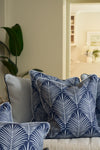 A cozy living room features a sofa with blue and white cushions, including the Oversized Mod Palm Pillow, next to a potted plant and illuminated by a bright doorway.