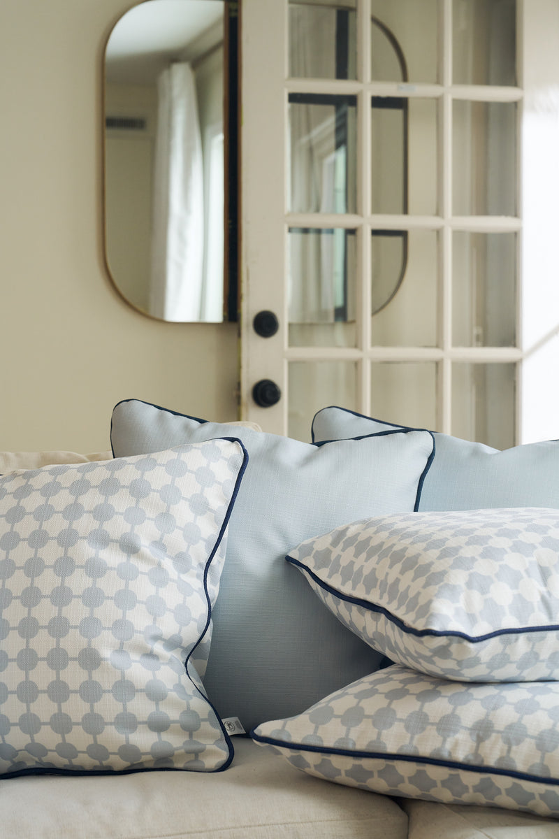 A close-up of a white sofa with four pillows, including the Oversized Mod Dot Grid Pillow with blue and white hand-drawn patterns, set before a glass-paneled door and a wall mirror reflecting curtains and light.