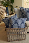 An Oversized Mod Palm Pillow with a blue and white leaf pattern sits in a wicker basket beside a beige sofa, lit candle, blue vase of flowers, and plants—perfect for cozy Southern-inspired decor.