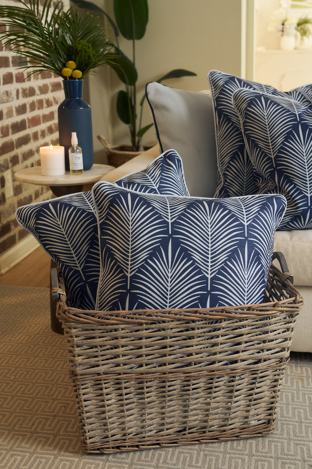 An Oversized Mod Palm Pillow with a blue and white leaf pattern sits in a wicker basket beside a beige sofa, lit candle, blue vase of flowers, and plants—perfect for cozy Southern-inspired decor.