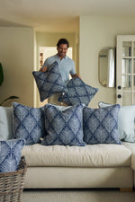A person smiles while arranging Oversized Mod Palm Pillows on a beige sofa in a cozy living room. Four matching blue and white pillows are on the sofa, with more nearby, enhancing the Southern-inspired decor.