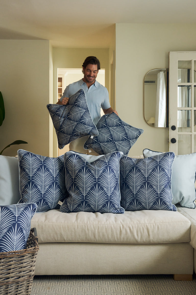 A person smiles while arranging Oversized Mod Palm Pillows on a beige sofa in a cozy living room. Four matching blue and white pillows are on the sofa, with more nearby, enhancing the Southern-inspired decor.