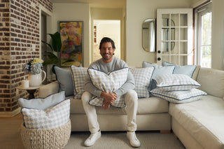 A man sits smiling on a beige sofa holding a checkered pillow, surrounded by more blue and white pillows. The cozy living room has a brick accent wall, plants, art, and natural light coming through glass doors.
