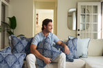 A brown-haired man sits on a sofa, smiling and holding an Oversized Mod Palm Pillow with a white leaf pattern. The sofa has matching pillows for a Southern charm, while a glass door and indoor plant complete the cozy scene.