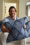 A brown-haired man with a beard smiles on a couch, holding the Oversized Mod Palm Pillow. Its blue cover and white palm print bring Southern charm to the softly lit, neutral-toned room.