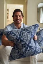 A brown-haired man with a beard smiles on a couch, holding the Oversized Mod Palm Pillow. Its blue cover and white palm print bring Southern charm to the softly lit, neutral-toned room.