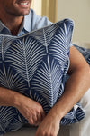 A man, wearing a light blue shirt with only his lower face and arms visible, smiles while holding the Oversized Mod Palm Pillow in navy blue with a white palm leaf design—an ideal accent for Southern-inspired decor.