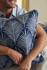 A man, wearing a light blue shirt with only his lower face and arms visible, smiles while holding the Oversized Mod Palm Pillow in navy blue with a white palm leaf design—an ideal accent for Southern-inspired decor.