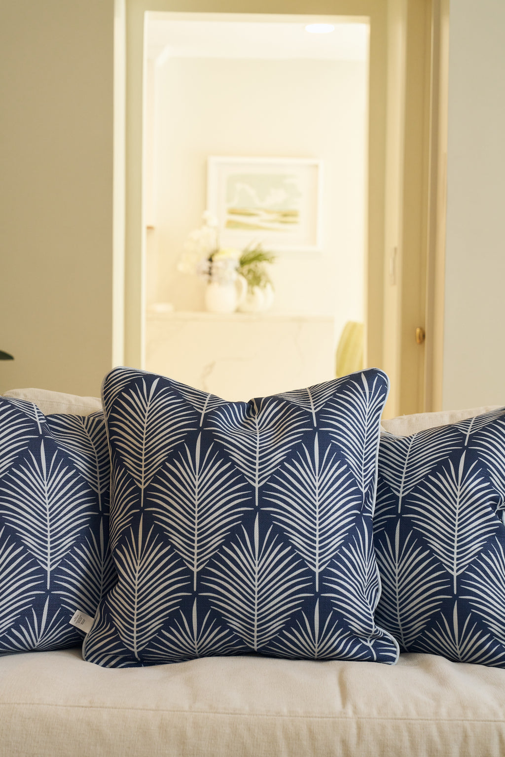 Three Oversized Mod Palm Pillows in navy blue with white leaf patterns are placed on a light sofa. Through the doorway, a bright room featuring Southern-style decor can be seen in the background.