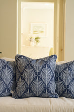 Three Oversized Mod Palm Pillows in navy blue with white leaf patterns are placed on a light sofa. Through the doorway, a bright room featuring Southern-style decor can be seen in the background.