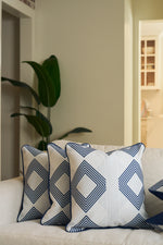 Two white throw pillows, one featuring the Mod Geo Diamond in Navy Pillow, sit on a light sofa. A leafy green plant is visible in the background, and an open doorway reveals a modern kitchen area.