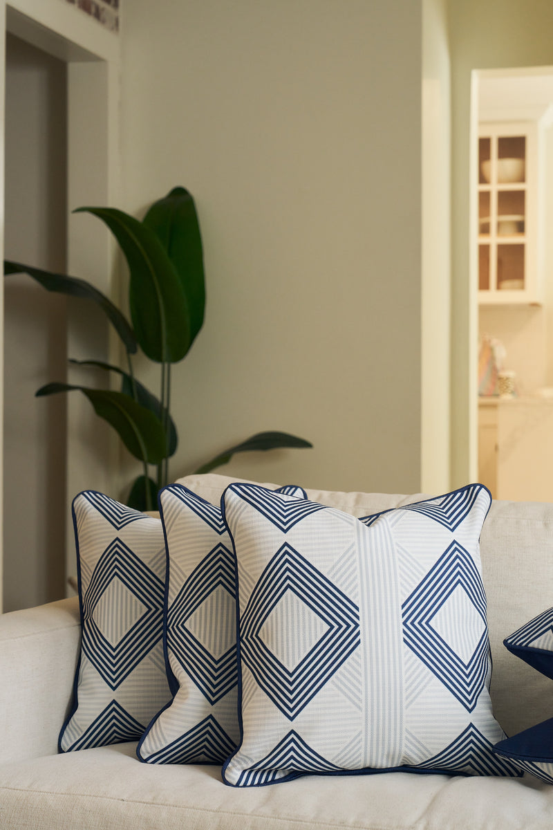 Two white throw pillows, one featuring the Mod Geo Diamond in Navy Pillow, sit on a light sofa. A leafy green plant is visible in the background, and an open doorway reveals a modern kitchen area.