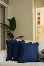 A Mod Geo Diamond in Navy Pillow and two other navy blue throw pillows are arranged on a light beige sofa in a cozy, modern living room, accented by a green potted plant and gentle natural light.