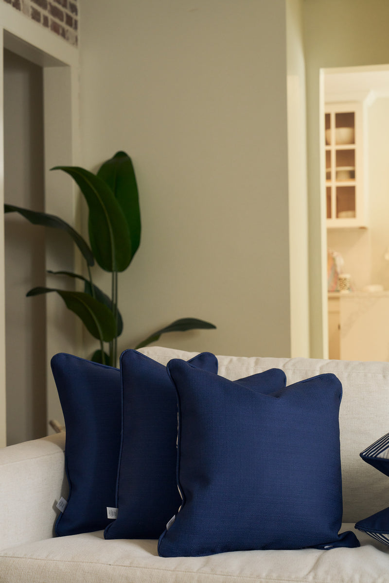 A Mod Geo Diamond in Navy Pillow and two other navy blue throw pillows are arranged on a light beige sofa in a cozy, modern living room, accented by a green potted plant and gentle natural light.