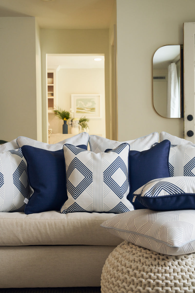 A beige sofa features a Mod Geo Diamond in Navy Pillow and blue patterned throws in a cozy living room. A round knit pouf sits in front, with modern decor and a mirror visible in the background.