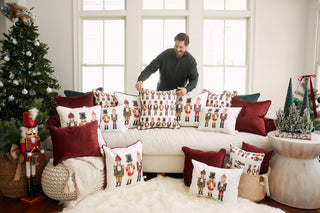 A man arranges the Nutcracker Line Up Pillow on a white sofa with deep red velvet pillows in a bright living room, while a decorated Christmas tree and festive decor add to the cozy holiday atmosphere.