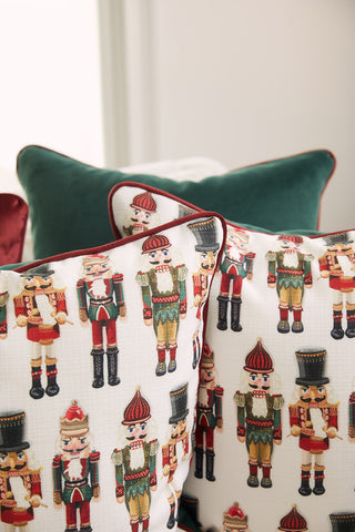 Two Nutcracker Line Up Pillows with a festive soldier design sit in front of a green velvet pillow with red piping, all arranged on a couch in a bright room.