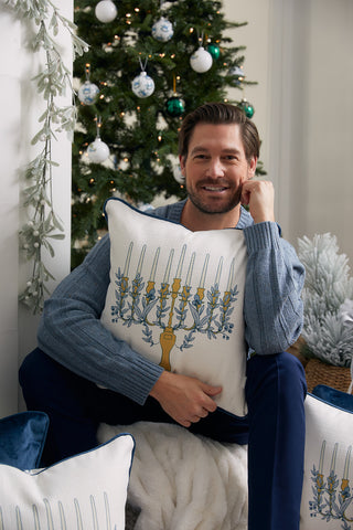A man smiles by a decorated Christmas tree, holding the Floral Hanukkah Pillow with a menorah print. Ornaments and festive greenery in the background highlight the harmonious blend of holiday traditions.
