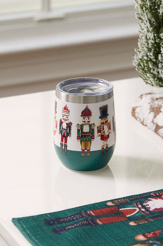 A Nutcracker Wine Tumbler featuring holiday designs is on a white table next to a small snowy artificial tree and a green festive placemat, with a window and beige siding in the background.