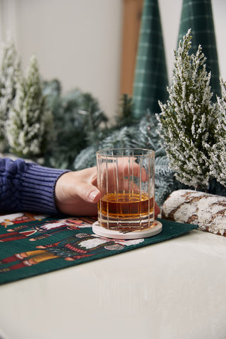 A hand in a blue sweater holds a glass of whiskey on a coaster, set atop festive holiday decor and snow-covered greenery, with drinks resting on the Reversible Nutcracker Placemat, Set of 4.