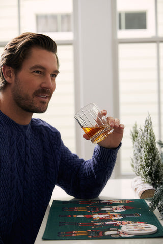 A man in a blue sweater sits at a table holding a glass of brown liquid, with festive holiday décor including the Reversible Nutcracker Placemat, Set of 4, and a small frosted decorative tree. Bright windows illuminate the background.