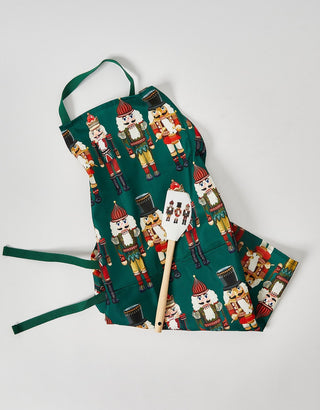 The Nutcracker Apron + Spatula Bundle features colorful soldier patterns on a white apron with a matching spatula, making it the ideal festive kitchen gift for holiday baking fans.