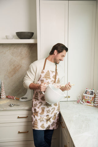 A man wearing a festive apron holds a Pink Gingerbread Spatula and mixing bowl, smiling in a kitchen filled with holiday treats like a gingerbread house and Christmas trees.