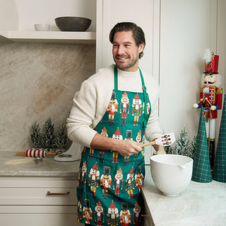 A man wearing a festive green apron with a repeating nutcracker pattern stands in a bright kitchen, holding a wooden spoon over a white bowl.