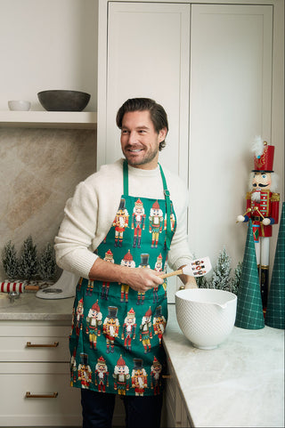 A man wearing the Nutcracker Apron + Spatula Bundle smiles while mixing ingredients in a festive kitchen with nutcracker decor—an ideal scene for discovering the perfect kitchen gift.