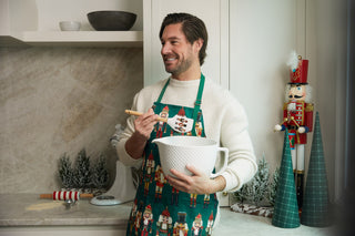 A smiling man in a Nutcracker Apron + Spatula Bundle stands in a festive kitchen, holding the spatula and ready for holiday baking. Nutcracker figurines and mini Christmas trees on the counter complete this perfect kitchen gift scene.