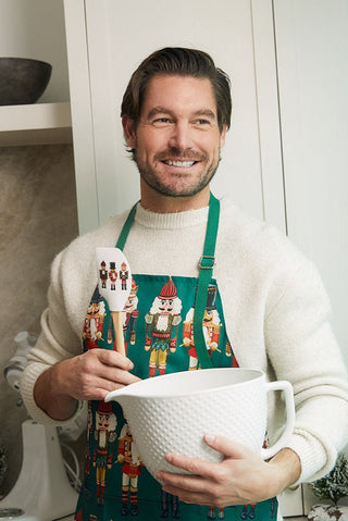 A smiling man in a festive nutcracker apron holds a white mixing bowl and the Nutcracker Spatula, made from BPA-free silicone—an ideal holiday baking gift—in his kitchen.
