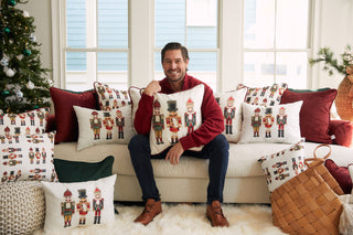 A smiling man in a red sweater sits on a couch holding the Nutcracker Holiday Pillow, surrounded by festive decor, velvet wine pillows, and a Christmas tree in a cozy living room with cheerful seasonal accents.