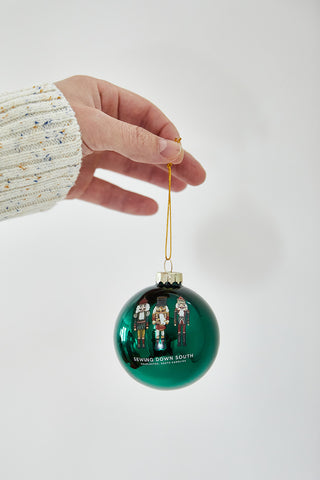 A hand in a cream sweater holds a shiny green Nutcracker Globe Ornament, featuring cartoon figures and SEWING DOWN SOUTH text, against a plain white backdrop. Available as single, 2 or 4 pack.