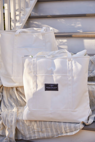 Two large SDS Label Logo Canvas Tote bags rest on striped fabric atop outdoor white steps. One features a front pocket, while soft sunlight casts gentle shadows across the scene.