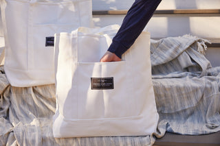 A person’s hand reaches into the front pocket of an SDS Label Logo Canvas Tote, resting on a striped blanket. Another large canvas tote bag sits nearby.