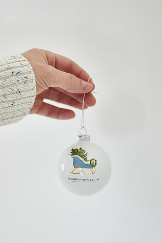 A hand in a cream knit sweater holds a Sleigh Globe Ornament—a white holiday ornament featuring an illustrated dog with a wreath and “Sewing Down South”—perfect for fans of unique festive décor. Available in single, 2 or 4 packs.