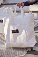 A person carries a large white SEW NEW: SDS Label Logo Canvas Tote, partially filled, outdoors against a striped fabric surface.