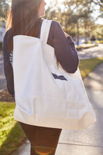 A person in a dark shirt carries the large SEW NEW: SDS Label Logo Canvas Tote with reinforced handles. The sun shines brightly, casting a warm glow on the tree-lined street.