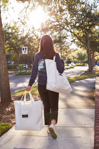A woman walks on a sunlit sidewalk carrying two SDS Label Logo Canvas Tote bags. She's dressed in a dark sweater, black pants, and green shoes, with trees and greenery lining the street behind her.