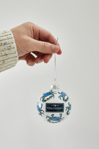 A hand in a cream and blue sweater holds a clear Crab Craze Globe Ornament featuring blue crabs and a Sewing Down South label—perfect for coastal holiday decor—against a plain light background. Available in single, 2, or 4 packs.