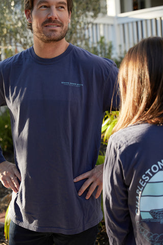 A man and woman stand outside a house wearing navy Charleston Pineapple Long Sleeve Tees. His shirt features text on the chest, while hers displays a back graphic reminiscent of the iconic Charleston Pineapple Fountain. They appear engaged in an animated conversation.