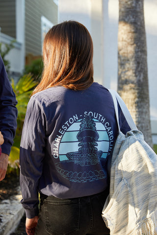 A person with long hair wears a navy Charleston Pineapple Long Sleeve Tee, featuring a Pineapple Fountain graphic and Charleston - South Carolina on the back, with a bag on their shoulder. Nearby, palm leaves and a tree trunk complete the scene.