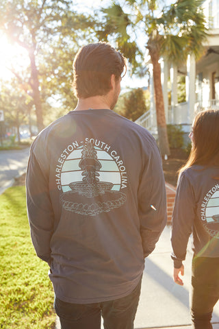 Two people walk down a sunny, palm-lined street, each wearing a Charleston Pineapple Long Sleeve Tee featuring Charleston, South Carolina and a Pineapple Fountain illustration on the back.