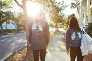 Two people walk down a sunlit street wearing matching navy Charleston Pineapple Long Sleeve Tees, one carrying a striped bag. Sunlight filters through the trees lining the sidewalk.