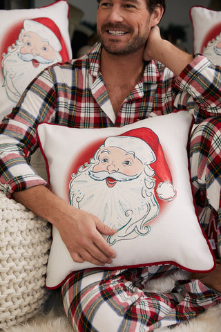 A smiling man in plaid pajamas holds a Vintage Stitch Santa Pillow from Sewing Down South, featuring a Santa Claus illustration and surrounded by other festive pillows.