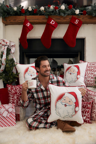 A smiling man in plaid pajamas sits by a fireplace, holding a Vintage Stitch Santa Pillow and a mug. The cozy space features festive garland, red stockings, Christmas pillows, and wrapped gifts for a charming holiday scene.