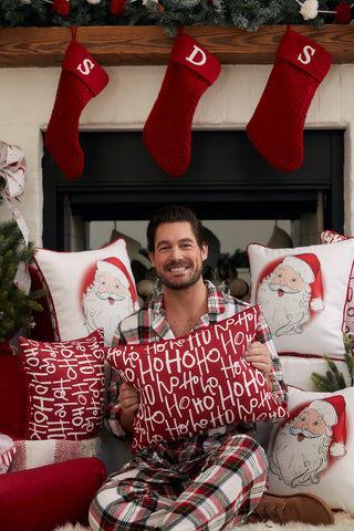 A man in plaid pajamas smiles by a fireplace with festive Santa pillows, three red stockings, and the Ho Ho Ho Lumbar Pillow—an ideal holiday accent.