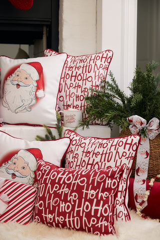 A cozy holiday scene with the Vintage Stitch Santa Pillow, featuring Santa faces and red-and-white Ho Ho Ho text, placed beside a basket of greenery and ribbon on a soft white rug.